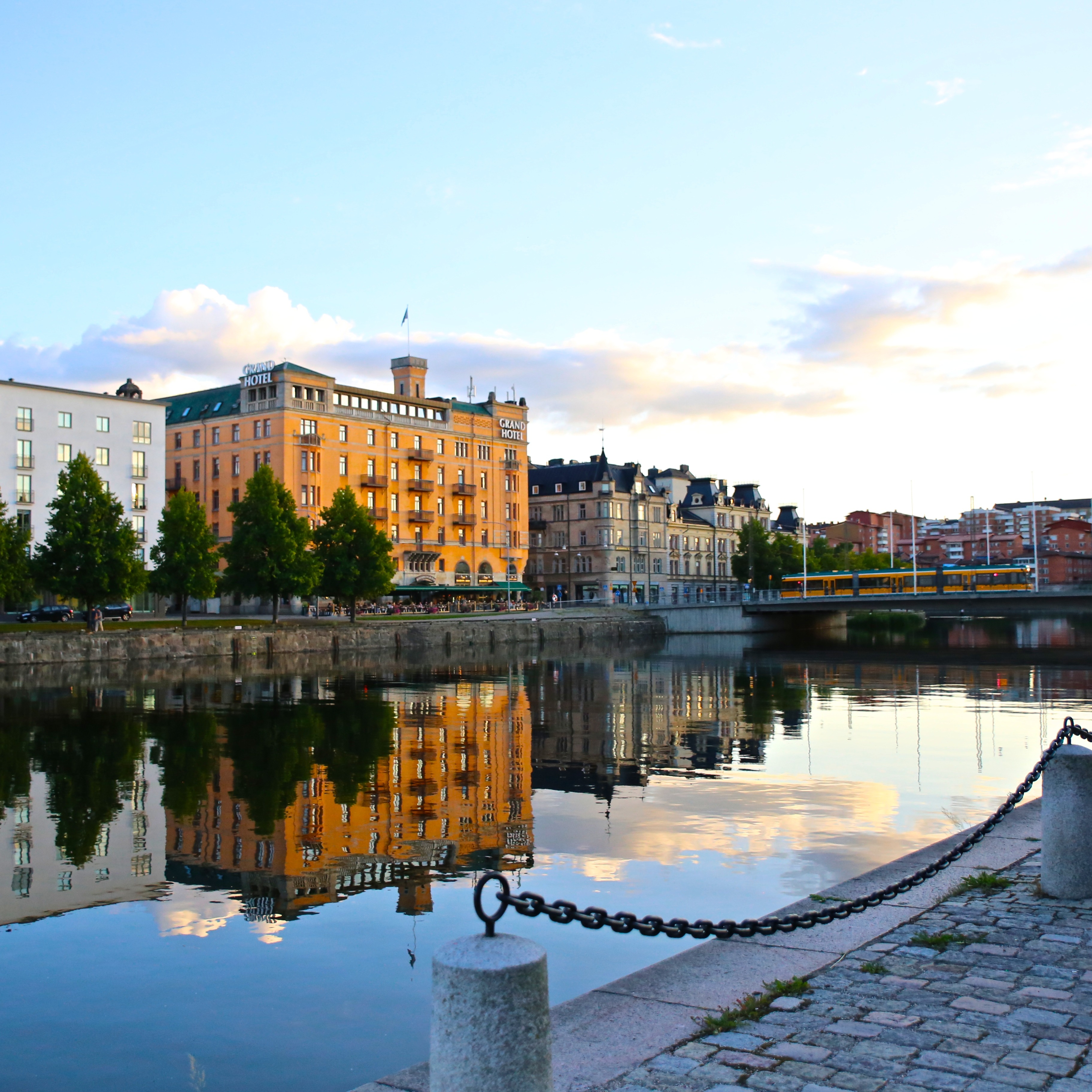 Cityscape of Norrköping in Sweden with buildings reflected in a calm river under a soft sky, a clock tower stands tall.