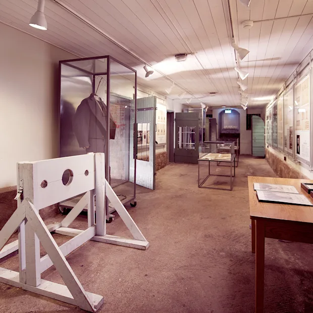A historical museum exhibit in a long hallway with a wooden stockade, display cases, and framed documents on the wall. Visible text: BRITTISK STRAFF.