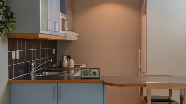 Ett litet kök med ljusblå skåp, en diskho och en bänkskiva med en radio och två vattenkokare. Ett matbord med stolar är delvis synligt. Beige väggar omger.
