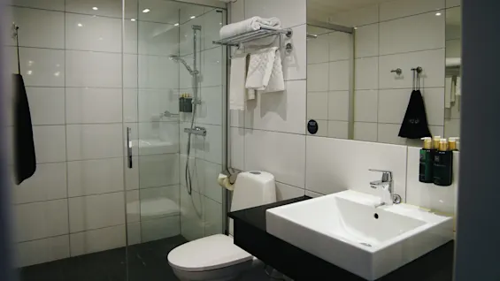 Badrum med en dusch med glasdörrar och toalettartiklar, en toalett och en vit handfat på en svart bänkskiva. Handdukar hänger på ställ, med vita kaklade väggar och taklampor.