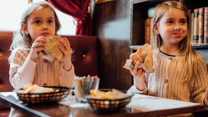 Two young children sit at a wooden table, each holding sandwiches. Colored pencils and baskets of fries are on the table. The cozy room has red curtains and a bookshelf.