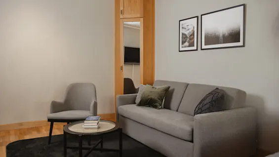 En grå sofa med to puter står ved siden av et lite rundt bord med bøker; en lenestol og innrammede bilder fullfører det moderne, minimalistiske rommet med tregulv.