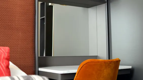 A vanity mirror stands on a desk, illuminated by an overhead lamp, in a room with a textured wall and a plush chair partially visible.