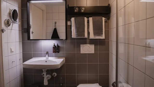 Ett badrum med en vit handfat under en upplyst spegel, flankeras av toalettartiklar. Handdukar hänger på en ställning ovanför toaletten. Grå och vita kakelplattor täcker väggarna.