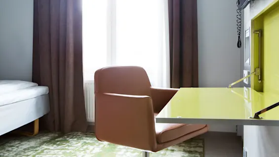 Brown chair sits by a green desk below a wall-mounted TV in a modern room with large window and brown curtains.