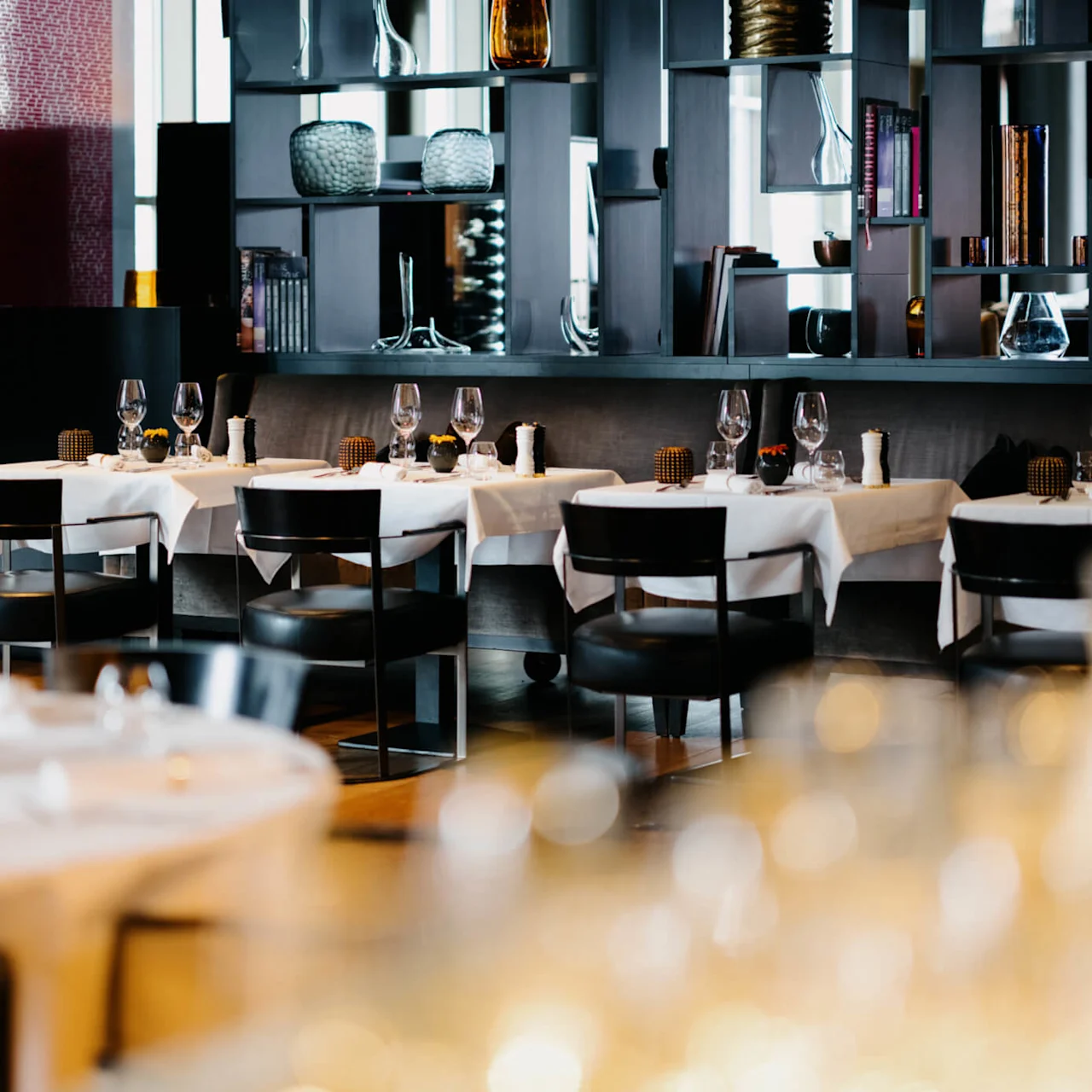 Bordene er dekket med hvite duker og vinglass, omgitt av svarte stoler i en stilig, moderne restaurantinteriør med dekorative hyller og dempet belysning.