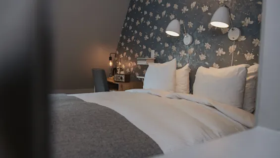 A neatly made bed with white pillows and a grey blanket sits in a cozy room. Two wall-mounted lights illuminate a floral-patterned accent wall, with a small wooden desk nearby.