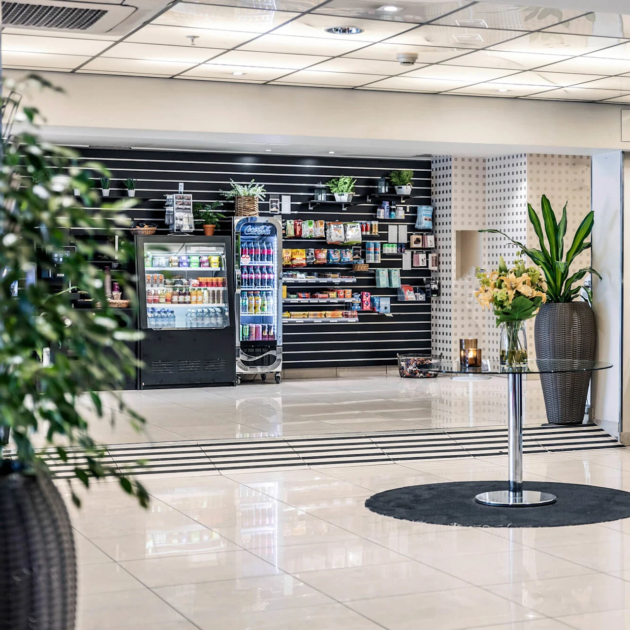 Reception-diskar står på kaklat golv; i närheten visas hyllor och kylar med snacks och drycker. Krukväxter ger grönska. Belyst tak förstärker modern lobby-atmosfär.