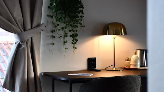 En mysig skrivbordshörna i ett rum med en varm lampa, en växt och ett fönster med gardiner. Perfekt för arbete eller avkoppling.