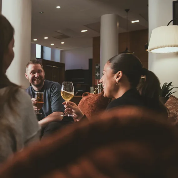 Tre personer sitter i en mysig lounge, njuter av drycker och ler under en konversation. En lampa ger varm belysning och växter ger en touch av grönska i bakgrunden.