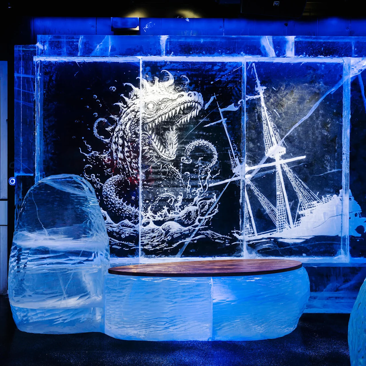 An ice sculpture depicts a fierce sea monster attacking a ship. The blue-lit environment enhances the chilling and dramatic scene, with smooth ice blocks forming seating in the foreground.
