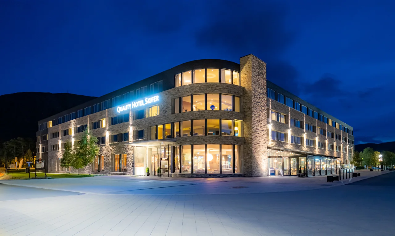 En stor hotellbygning med skiltet "Quality Hotel Sogndal" er opplyst om natten. Den står med moderne arkitektur i et åpent, brosteinsbelagt gårdsplass under en klar, dypblå himmel.