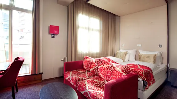 Et moderne hotellrom med en rød sovesofa med blomstermønster og balkongutsikt, opplyst av naturlig sollys.