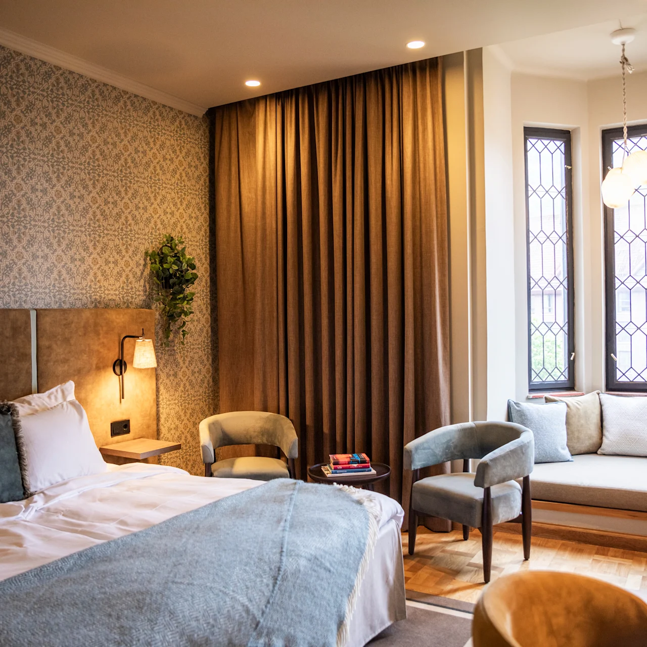 A cozy, neatly furnished bedroom features a large bed, a wall-mounted TV, and a sitting area by stained glass windows with cushions and books, creating a warm, inviting atmosphere.