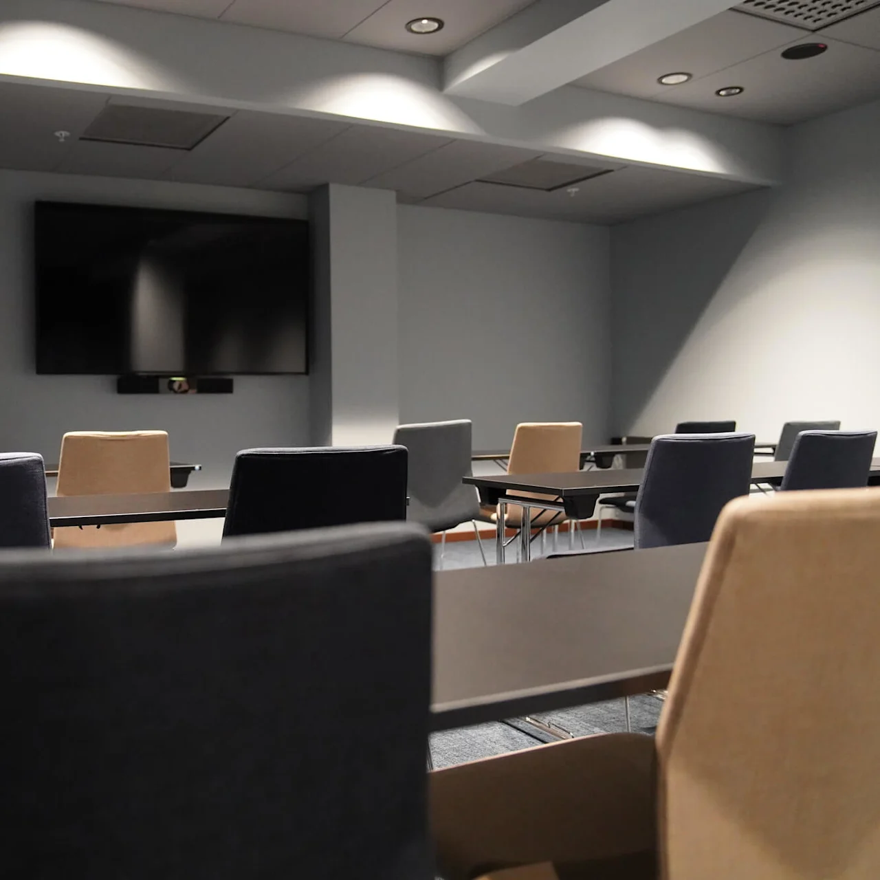 Rækker af tomme stole vender mod et vægmonteret TV i et moderne, svagt oplyst konferencelokale med grå vægge og loftslamper.