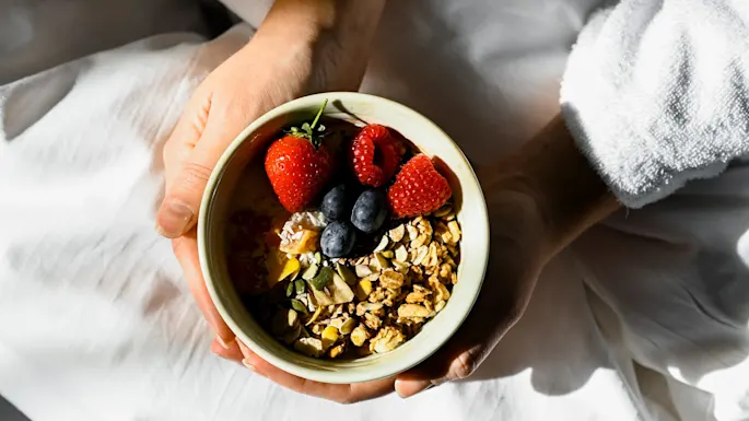 A bowl of granola with berries is held by two hands. The person is wearing a white robe and sitting on white bedding, with sunlight casting gentle shadows.