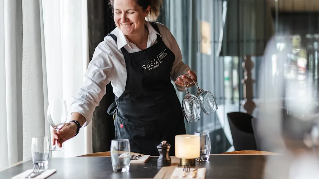 En servitør, iført et forkle merket "The Social", setter glass på et bord i en restaurant. Bordet er elegant dekket med bestikk, glass og et tent lys.