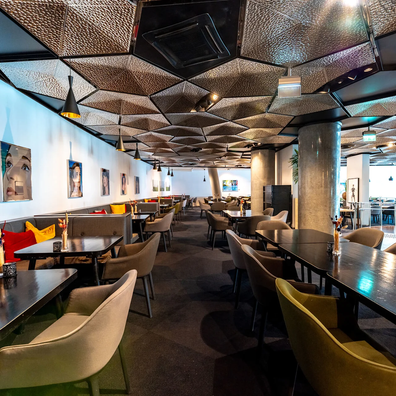 Spiseborde med stole står på række i en stilfuld, moderne restaurant med geometriske loftsmønstre og farverige puder. Kunstværker pryder væggene, hvilket forstærker den moderne atmosfære.
