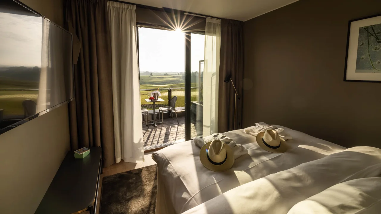 A hotel room with sunlight streaming through open glass doors onto a bed, leading to a balcony overlooking a green landscape at Lily Country Club in Norwegian Klofta.