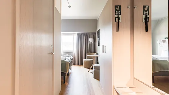 A bright and modern hotel room interior, featuring a light wood entrance area leading to a comfortable bed and a cozy seating space.