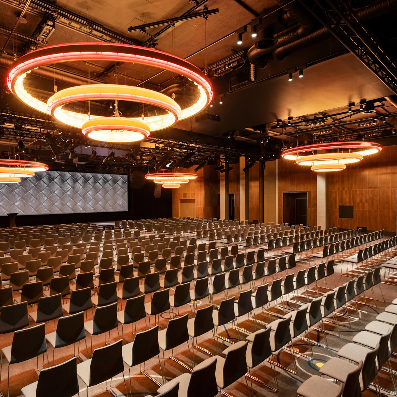 Rows of empty chairs face a large stage in an auditorium, illuminated by circular ceiling lights. The room features wooden walls and a carpeted floor.