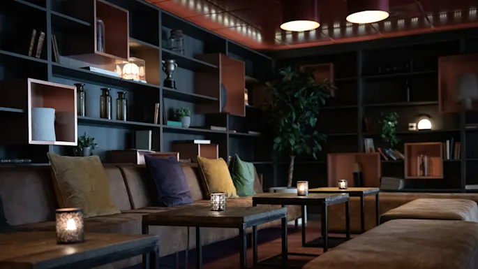 Stearinlysfat er plassert ved en brun sofa med fargerike puter. Bakgrunnen har hyller med bøker, planter og dekorative gjenstander, og skaper en koselig, dempet opplyst stuelignende atmosfære.
