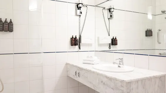 Siisti ja moderni hotellin kylpyhuone, jossa on marmorinen pesuallas, peili, seinään kiinnitetyt hygieniatuotteet ja hiustenkuivaaja, valmiina käyttöön.