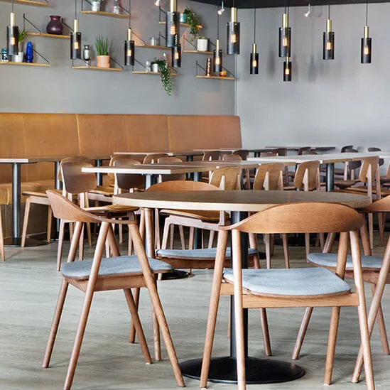 Træstole omgiver borde i en moderne café. Læderbås langs væggen, mens hylder holder planter og dekoration ovenover. Hængende lamper i loftet.