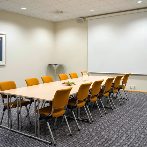Konferansebord med oransje stoler rundt det, vannmugger og frukt på toppen, i et rom med en stor tom skjerm og mønstret teppe.