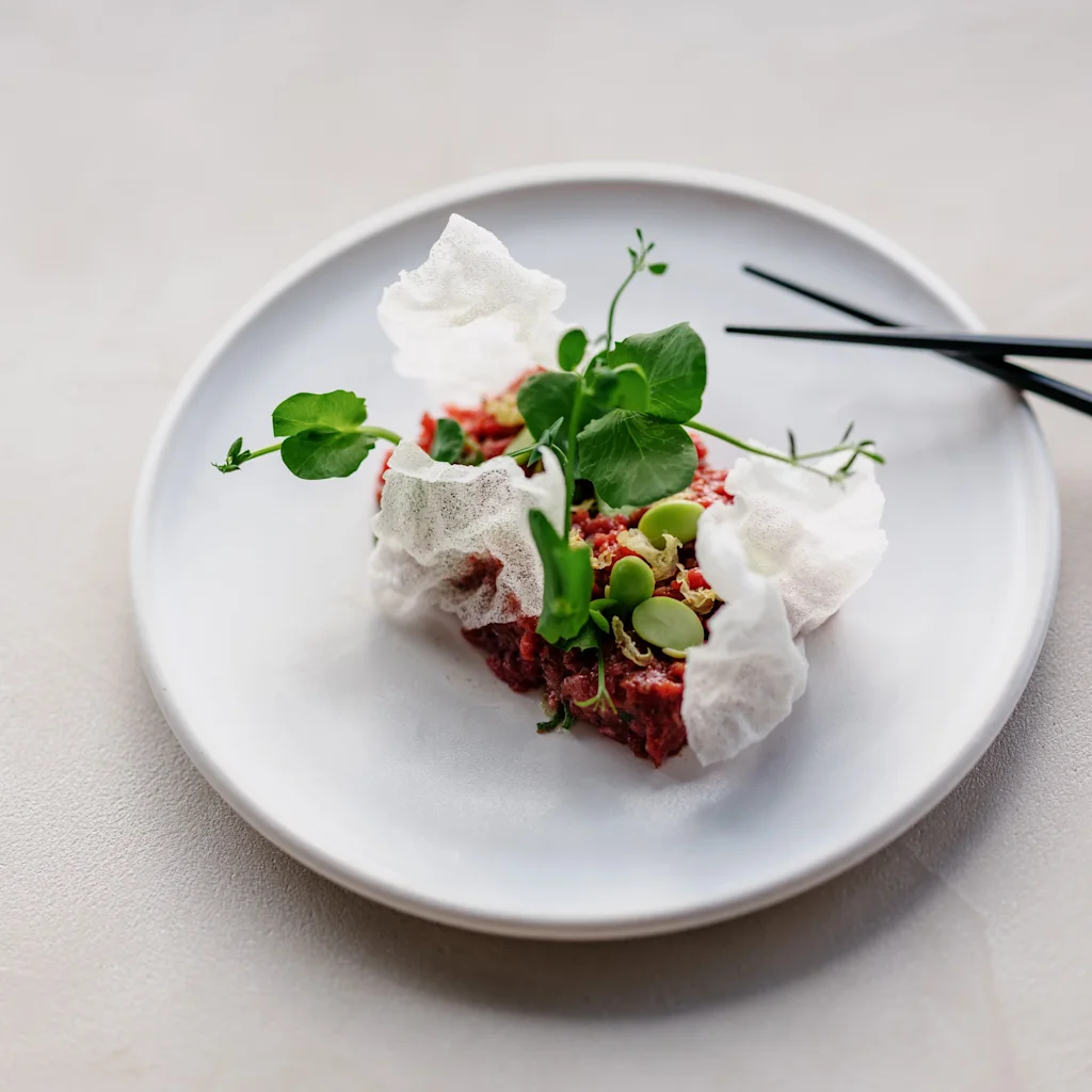 En gourmetrett med tartar, grønne garnityrer og sprø elementer, servert på en hvit tallerken med svarte spisepinner.