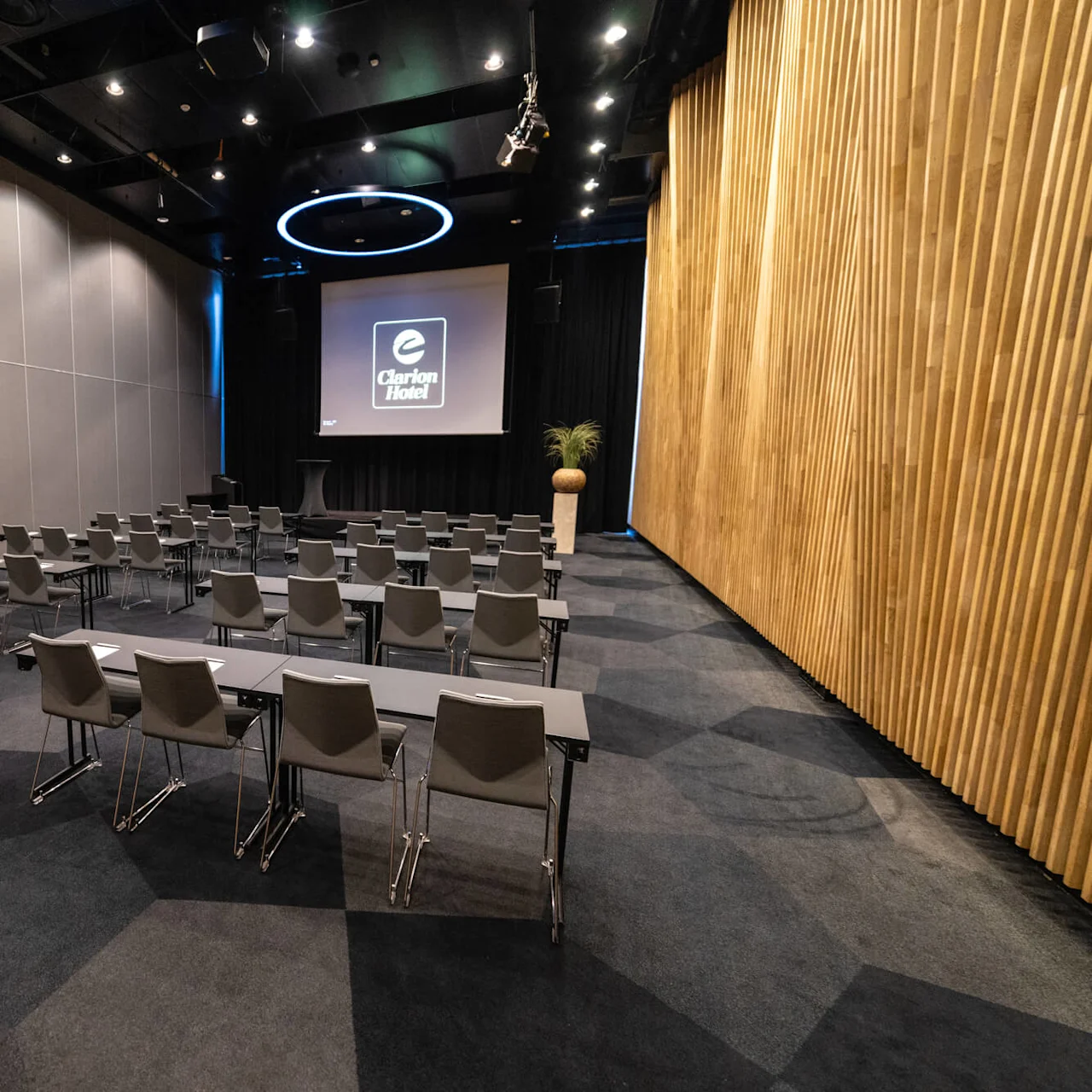 Stole og borde er arrangeret i rækker mod en projektorskærm med teksten "Clarion Hotel". Konferencelokalet har højt til loftet, trævægge og dæmpet belysning.