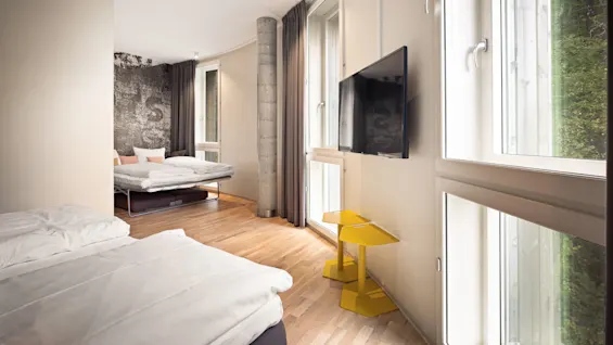 Two beds rest against a patterned wall, with large windows illuminating a minimalist room. A wall-mounted TV faces vibrant yellow tables, complemented by wooden flooring and neutral curtains.