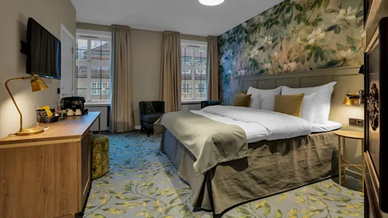 Ett modernt hotellrum med en säng täckt med vita lakan, omgivet av blommig tapet, gardiner, en TV, en lampa och utsikt över byggnader utanför.
