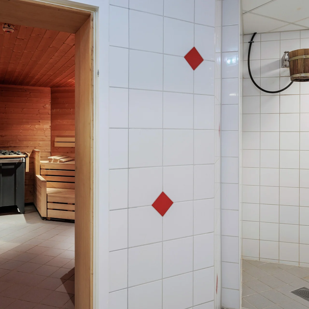 A wooden sauna with benches and a heater is on the left. A tiled room with a wooden water bucket suspended from the wall is on the right. 