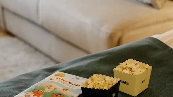 Two popcorn containers and a children's activity booklet, titled "Lollo & Bernie," rest on a grey blanket in a cozy living room with a beige sofa in the background. Text on popcorn containers: "pop! QUALITY HOTEL" and "make me".