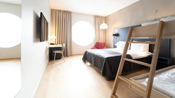 A hotel room with a bed, bunk bed, and desk with a chair beneath a wall-mounted TV. A large round window lets in light, illuminating beige curtains and wood flooring.