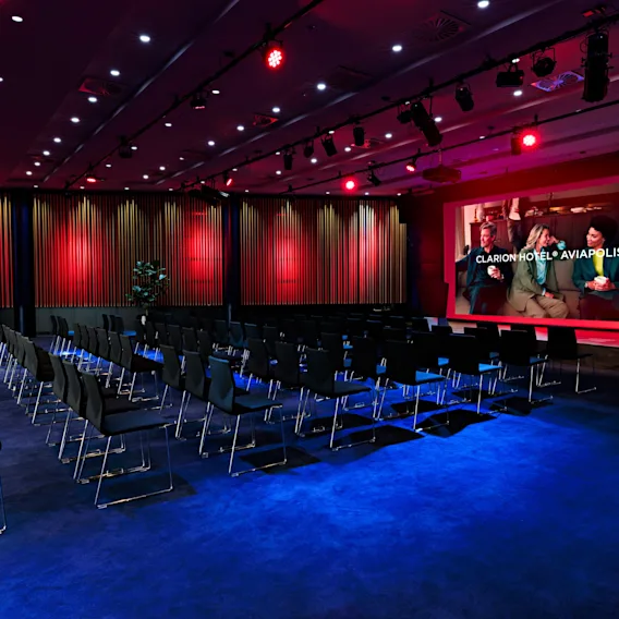 Konferenssihuone, jossa on riviä tuoleja suurta näyttöä kohti. Näytöllä näkyy "AWARDS" ja ihmisiä. Huoneessa on sininen matto ja punainen valaistus.