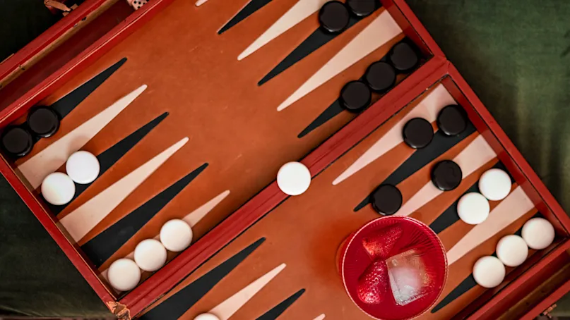 Backgammon board with white and black pieces arranged for a game, sits on a table. A red dice cup, filled with two dice, is nearby.
