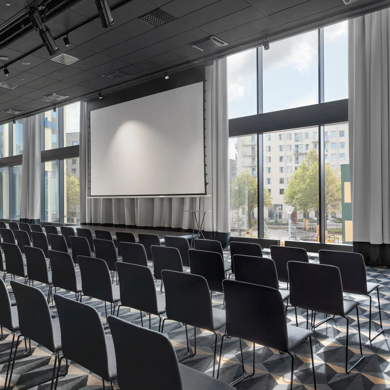 Rader av tomma svarta stolar vetter mot en stor projektorduk i ett rymligt, välbelyst konferensrum med fönster från golv till tak och ett mönstrat golv. Stadsbyggnader är synliga utanför.