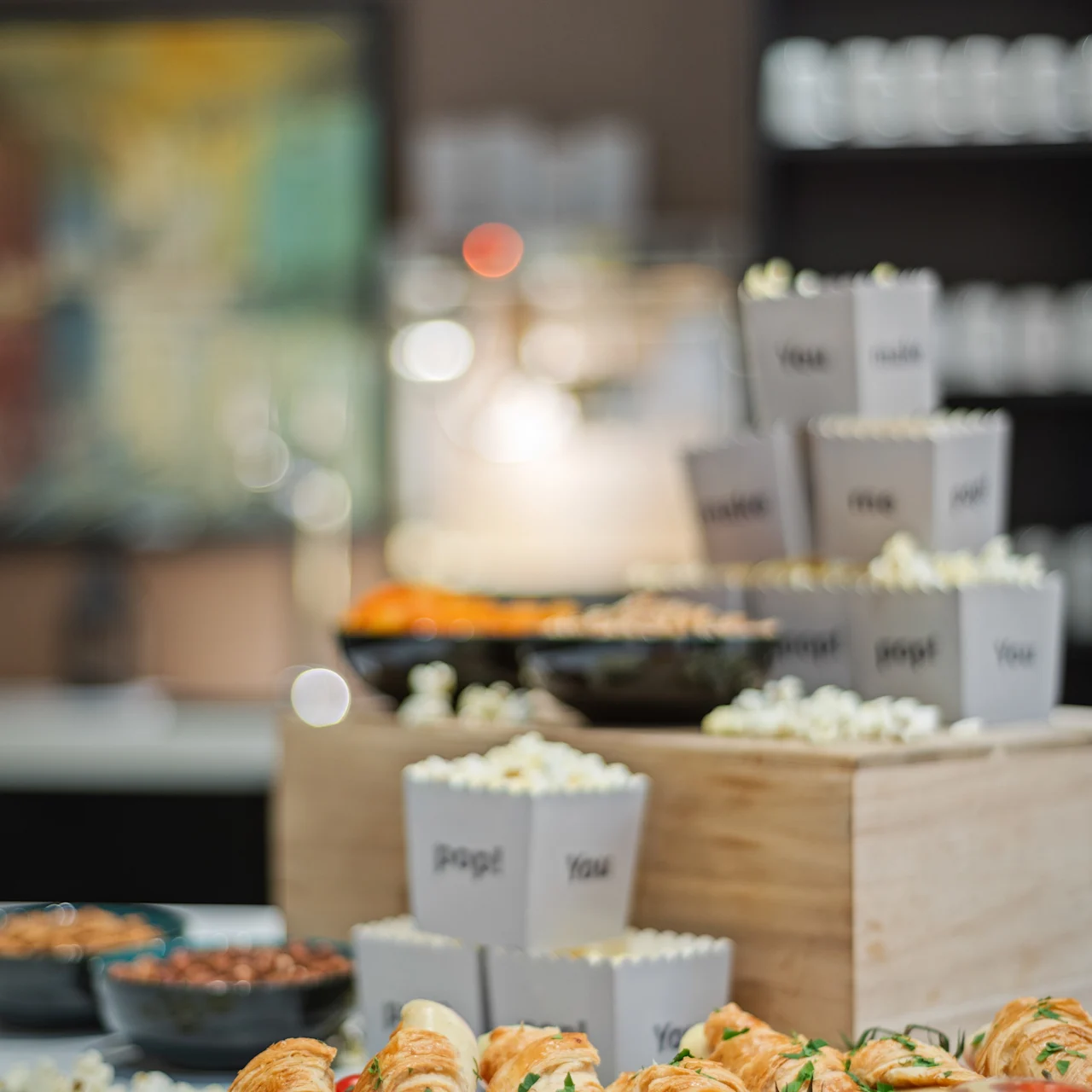 Bagværk fyldt med skinke og tomater ligger på et træbræt, omgivet af popcorn i æsker mærket "pop!" og "You." Miljøet er en afslappet buffet.