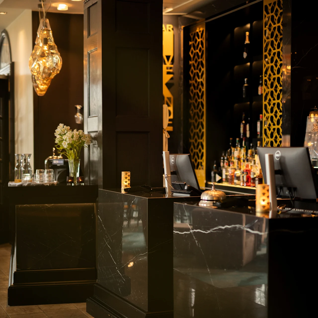 A modern bar counter, equipped with liquor bottles and computer screens, features elegant black marble surfaces, flanked by decorative patterned panels, softly lit in a stylish interior setting.