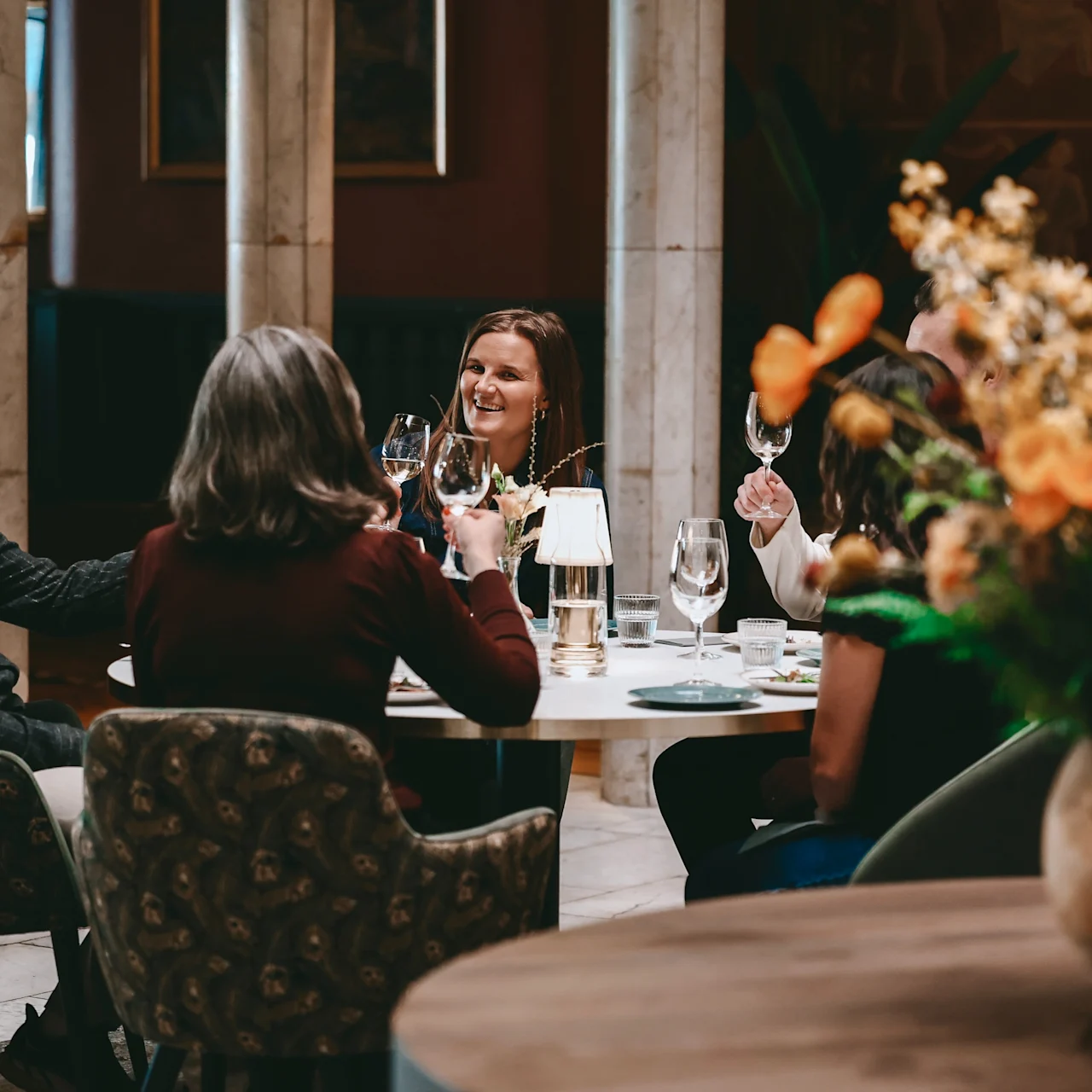 A group sits at a round table in a cozy restaurant, raising glasses in a toast. Warm lighting and orange flowers create a relaxed atmosphere.