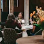 A group sits at a round table in a cozy restaurant, raising glasses in a toast. Warm lighting and orange flowers create a relaxed atmosphere.