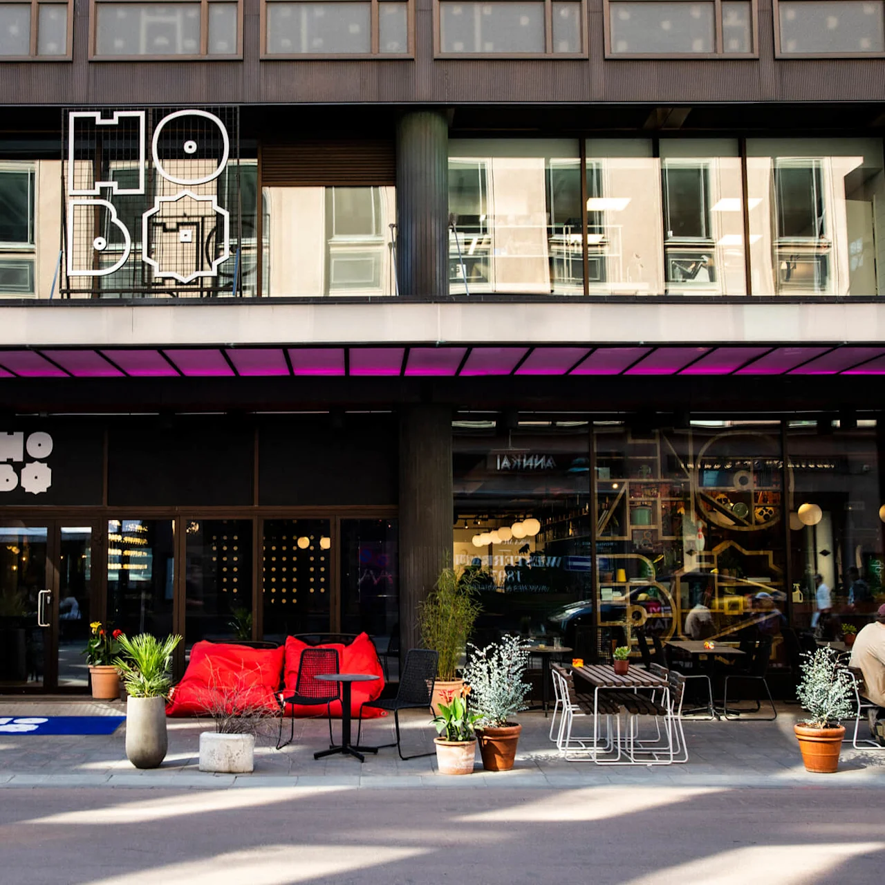  En moderne bygningsfacade med skiltet "HO BO". Rød siddeplads og potteplanter pryder udendørsområdet, hvor folk er samlet. En person går forbi.