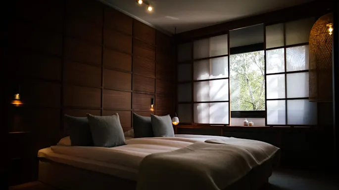 A bed with white linens in a dimly lit room with wooden walls and a large window showing greenery outside.