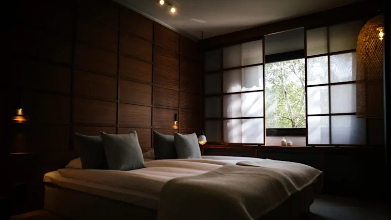 A bed with white linens in a dimly lit room with wooden walls and a large window showing greenery outside.