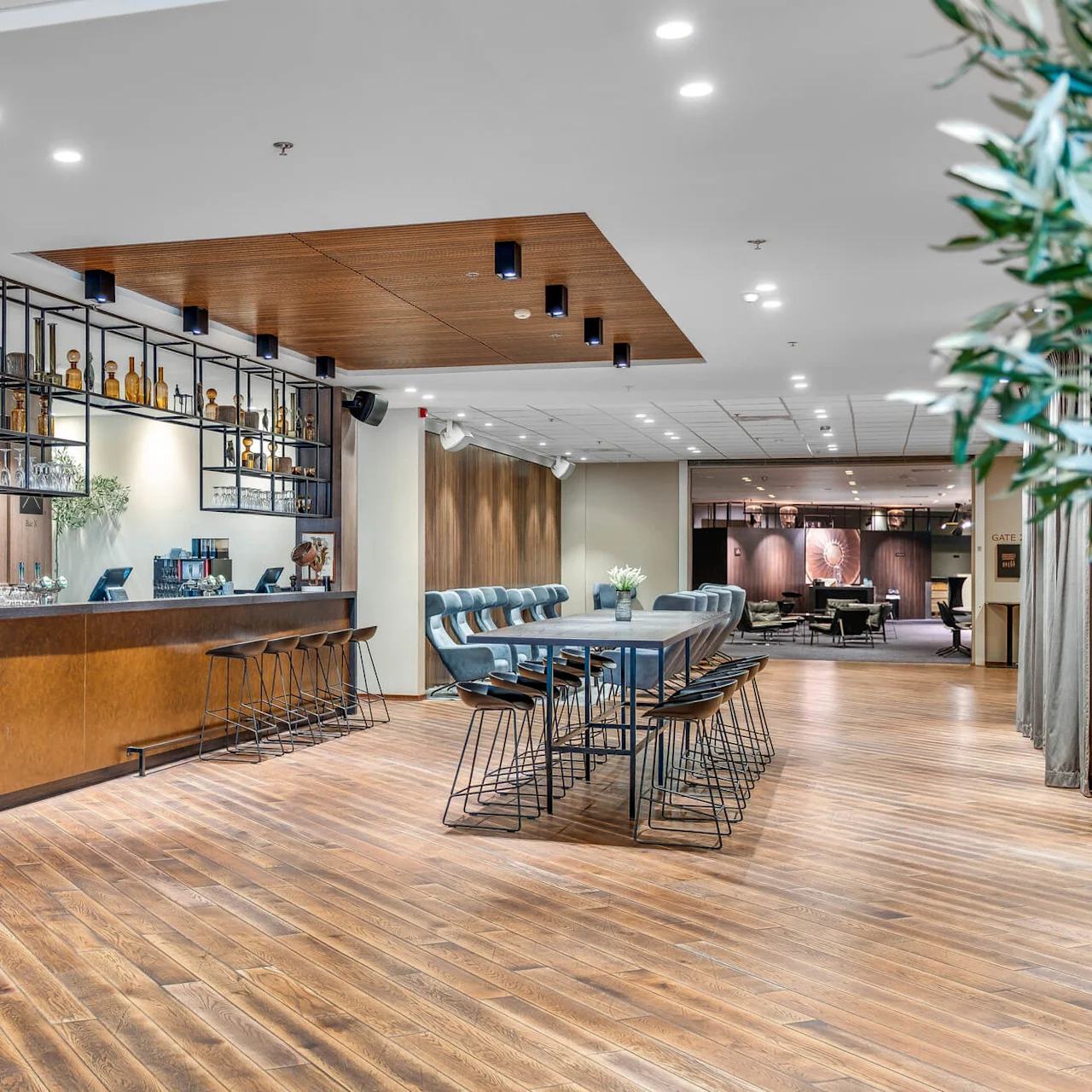 A modern hotel lobby with wooden floors, a coffee bar counter, stools, and tables; warm lighting creates a welcoming atmosphere.