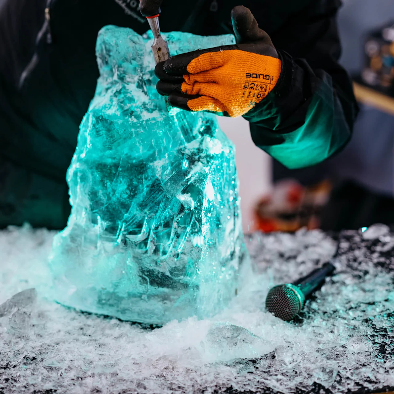A person in winter clothing carves a glowing ice block with a chisel, wearing orange gloves. Shaved ice surrounds them on a table with a microphone nearby.