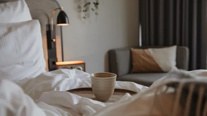 En kopp står på et trebrett oppå en seng med hvite dyner. En moderne lampe på et nattbord lyser mykt opp rommet, som har en sofa med puter.
