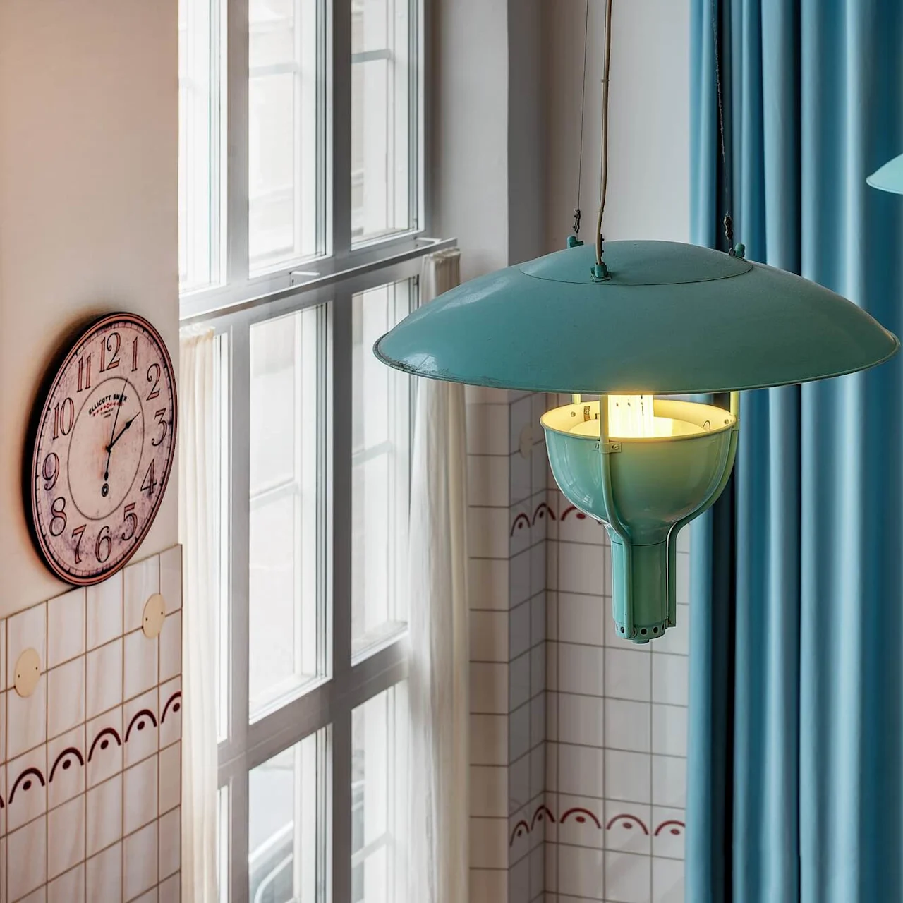 En väggklocka i vintagestil visar 10:10 nära ett draperat fönster, med två industriella hänglampor framför blå gardiner som ger varmt ljus.
